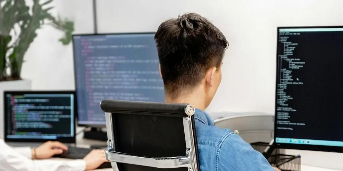 Ein Mann arbeitet an einem Schreibtisch mit mehreren Monitoren, auf denen Quelltext zu sehen ist